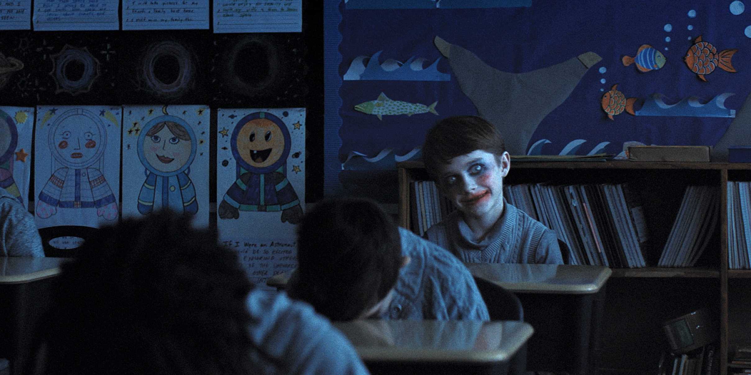 Алекс с широкой улыбкой и чрезмерным макияжем, когда одноклассники спят за партами в фильме "Weapons"