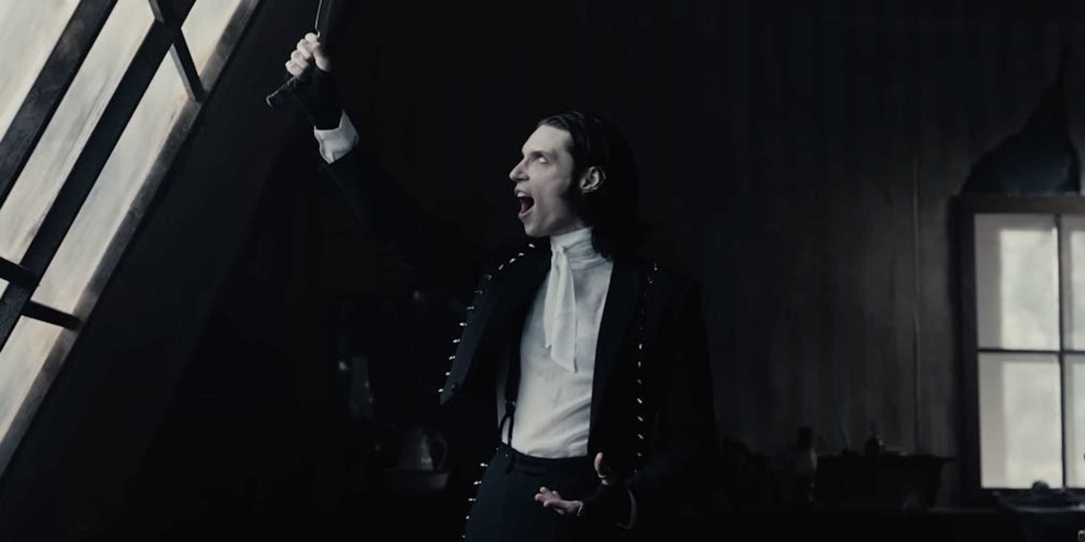 На заднем плане черные стены. Человек одет в черные брюки, черную куртку и белую футболку. У него также черные, уложенные назад волосы. Он держит бритву, направленную в окно, и кричит.