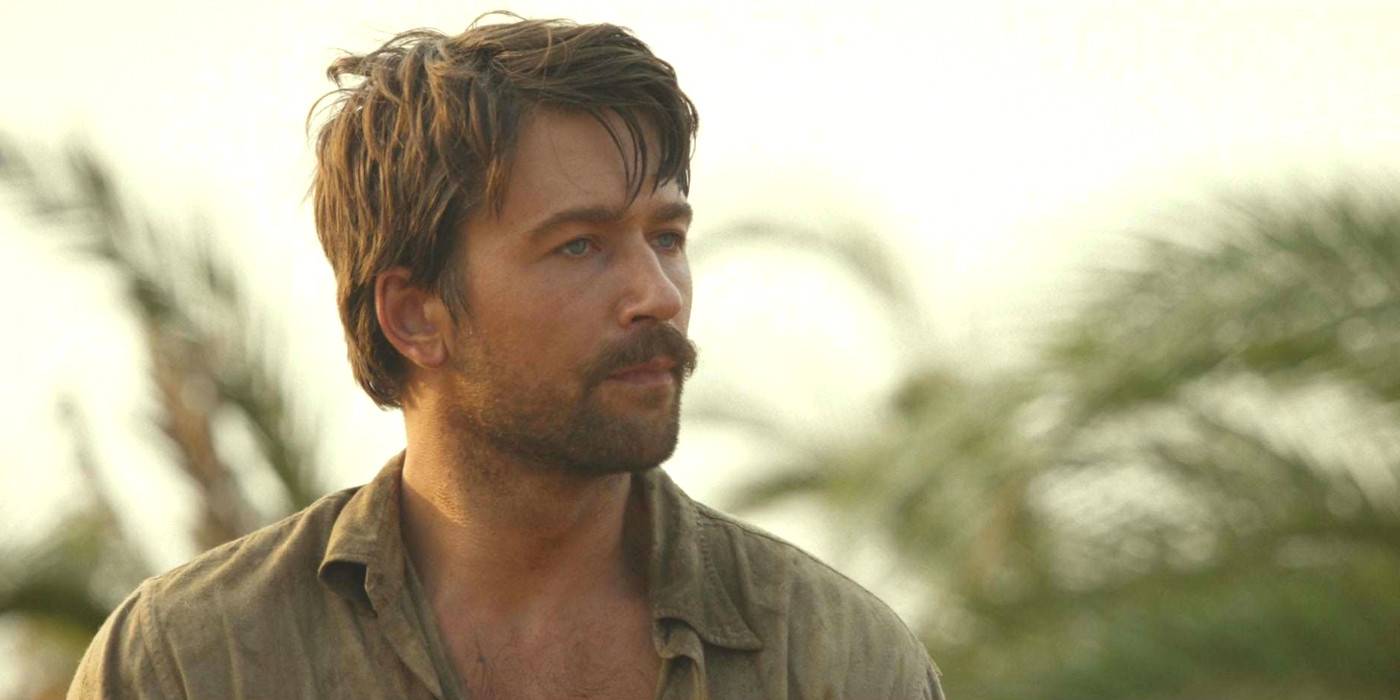 Брендон Склен в роли Спенсера Даттона в 1 сезоне сериала "1923" с растрепанными волосами и усами на фоне пальм