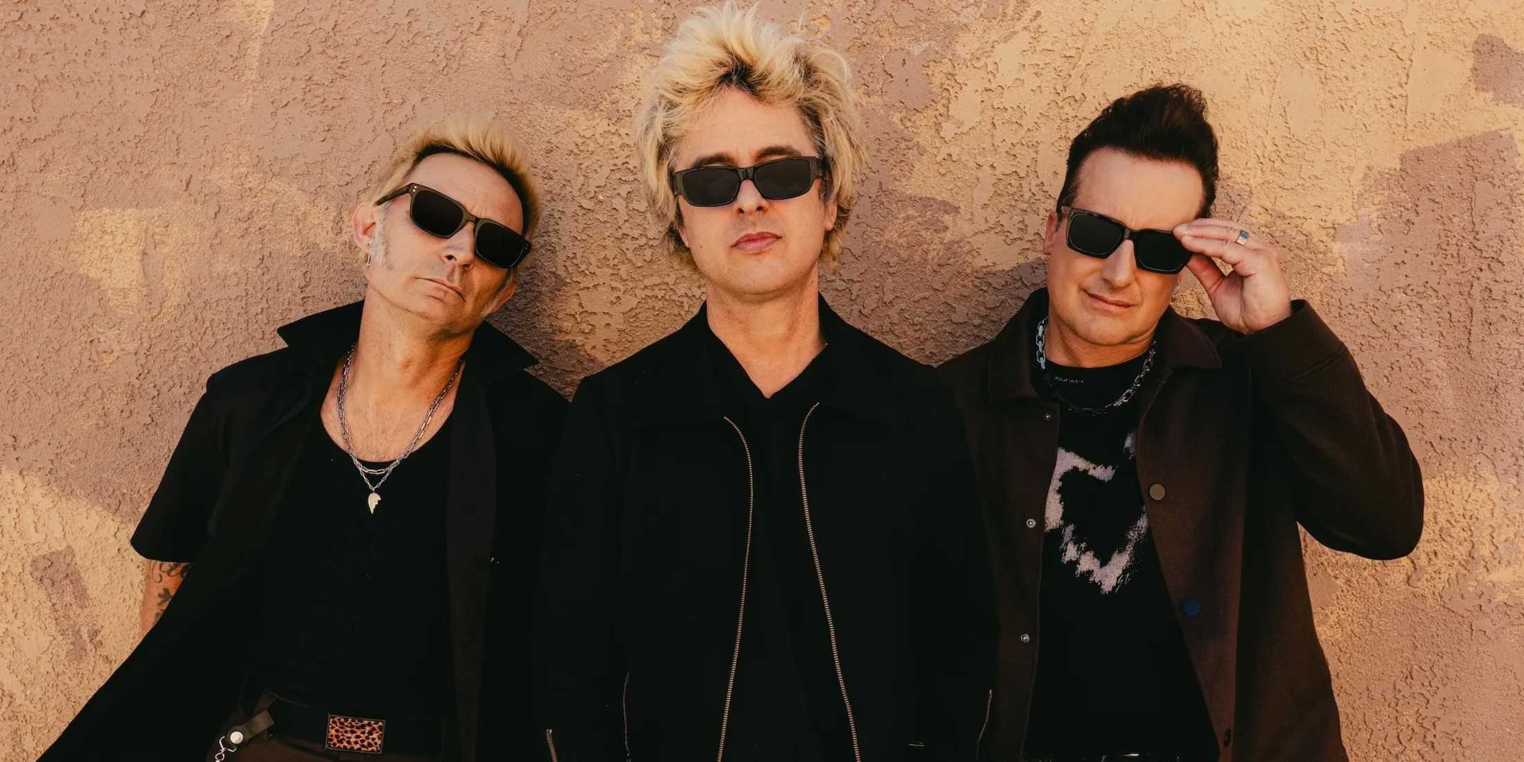 Три участника Green Day стоят перед белой стеной. У каждого на солнцезащитных очках и длинных куртках. 