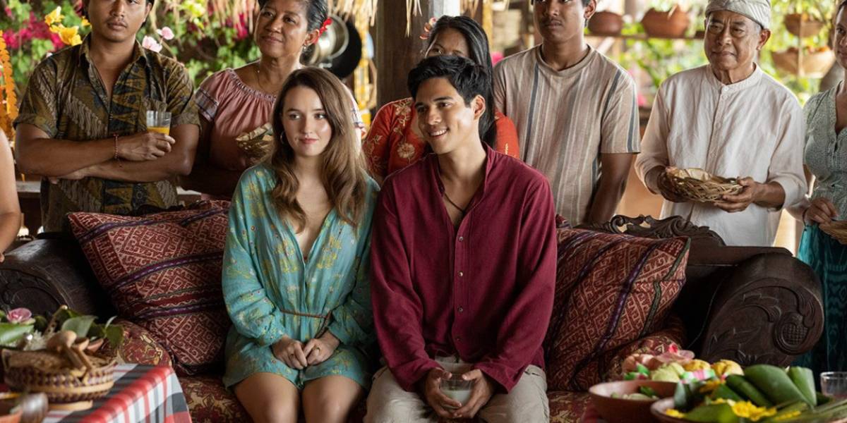 Кейтлин Девер и Макс Бутье, сидящие рядом, с местными жителями, стоящими за ними в фильме "Ticket To Paradise"