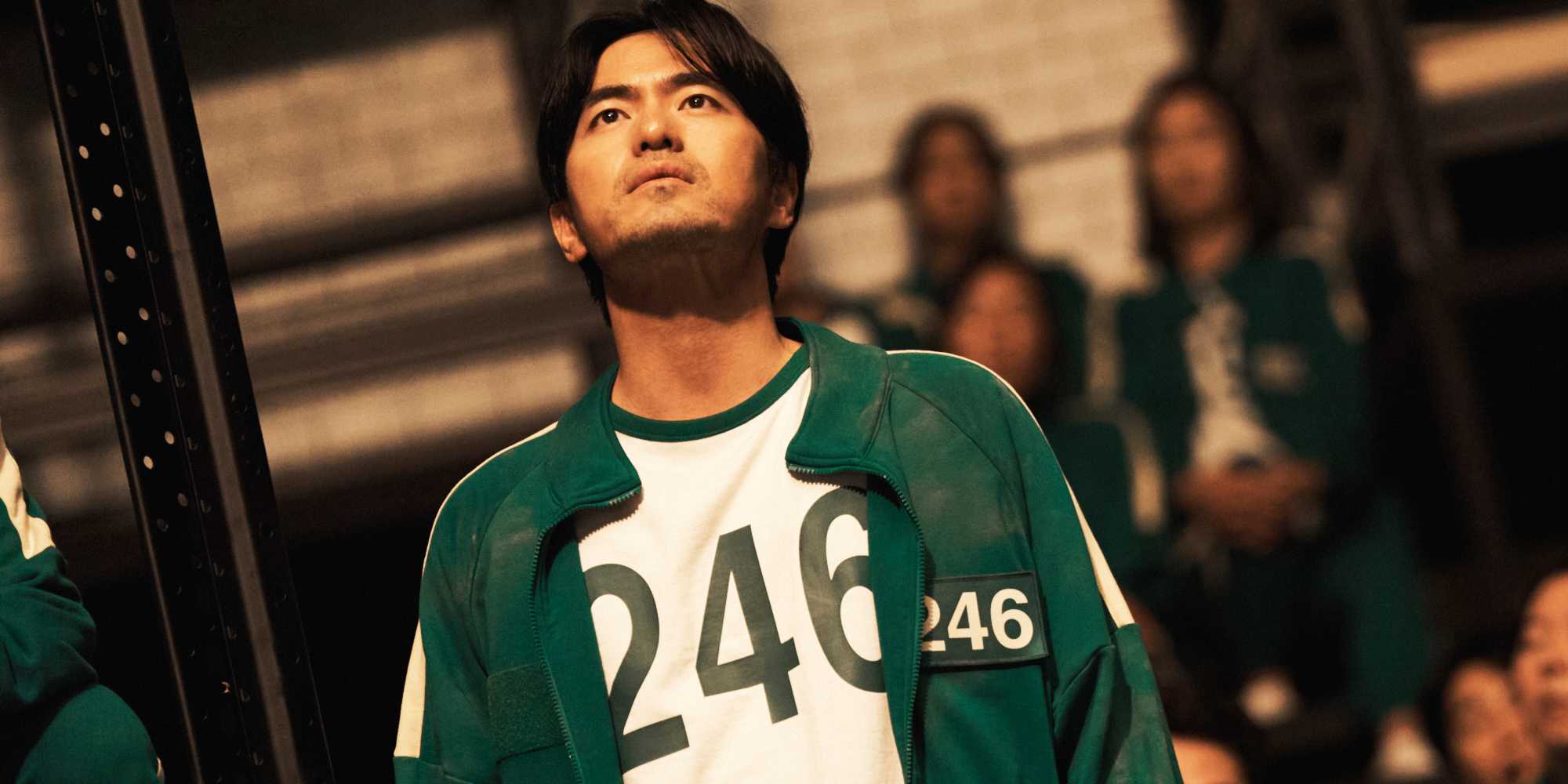 Гён-сок смотрит вверх, одетый в свою куртку игрока 246 в 2 сезоне "Игра в кальмара"