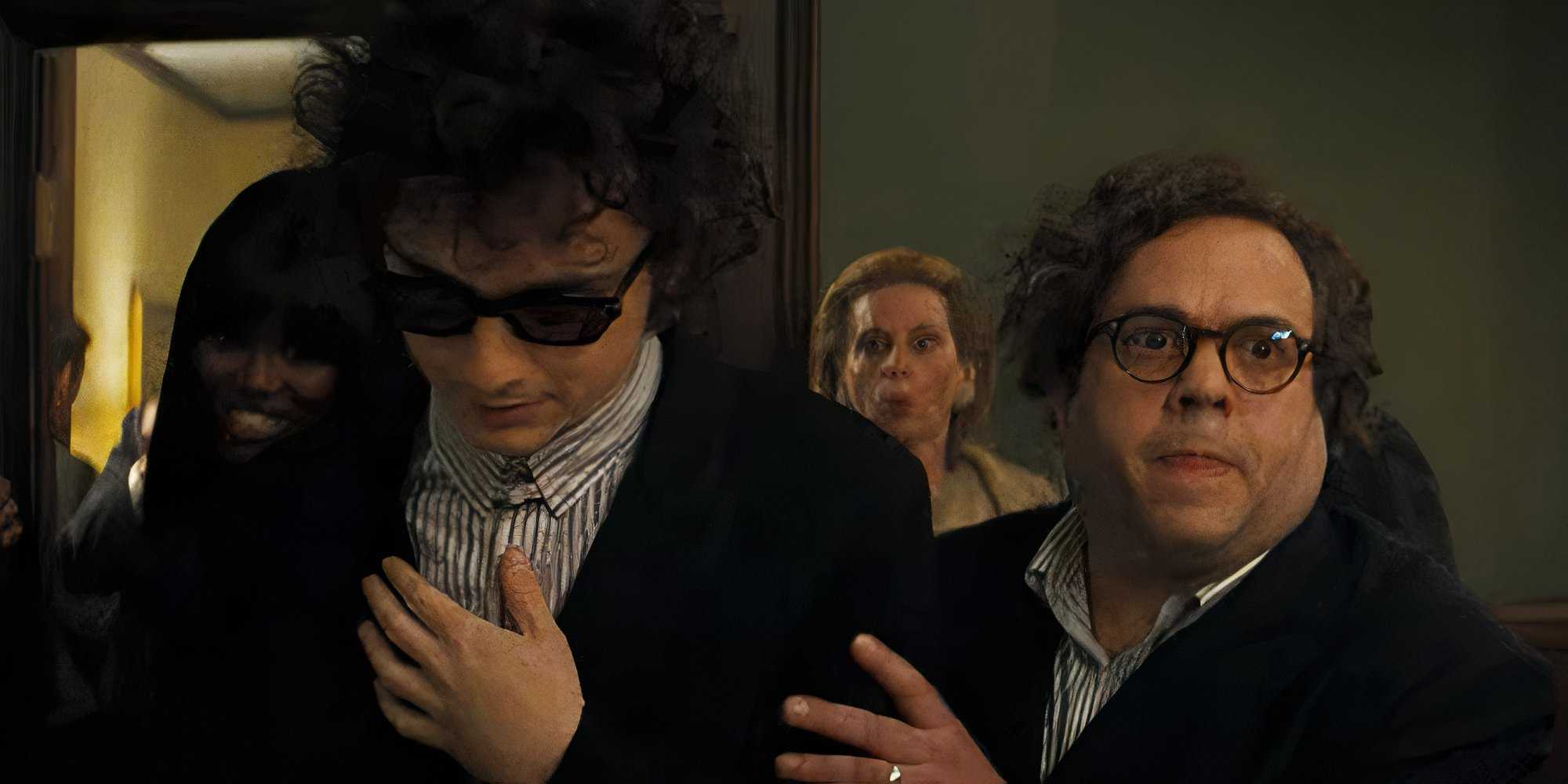 Боб Дилан (Тимоти Шаламе) под руководством своего менеджера, Альберта Гросмана (Дэн Фоглер), проходит через переполненную комнату, нося солнцезащитные очки и глядя вниз.