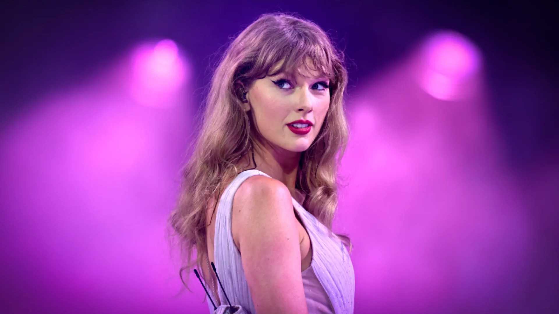 Taylor Swift позирует перед фиолетовыми прожекторами с невыразительной улыбкой.