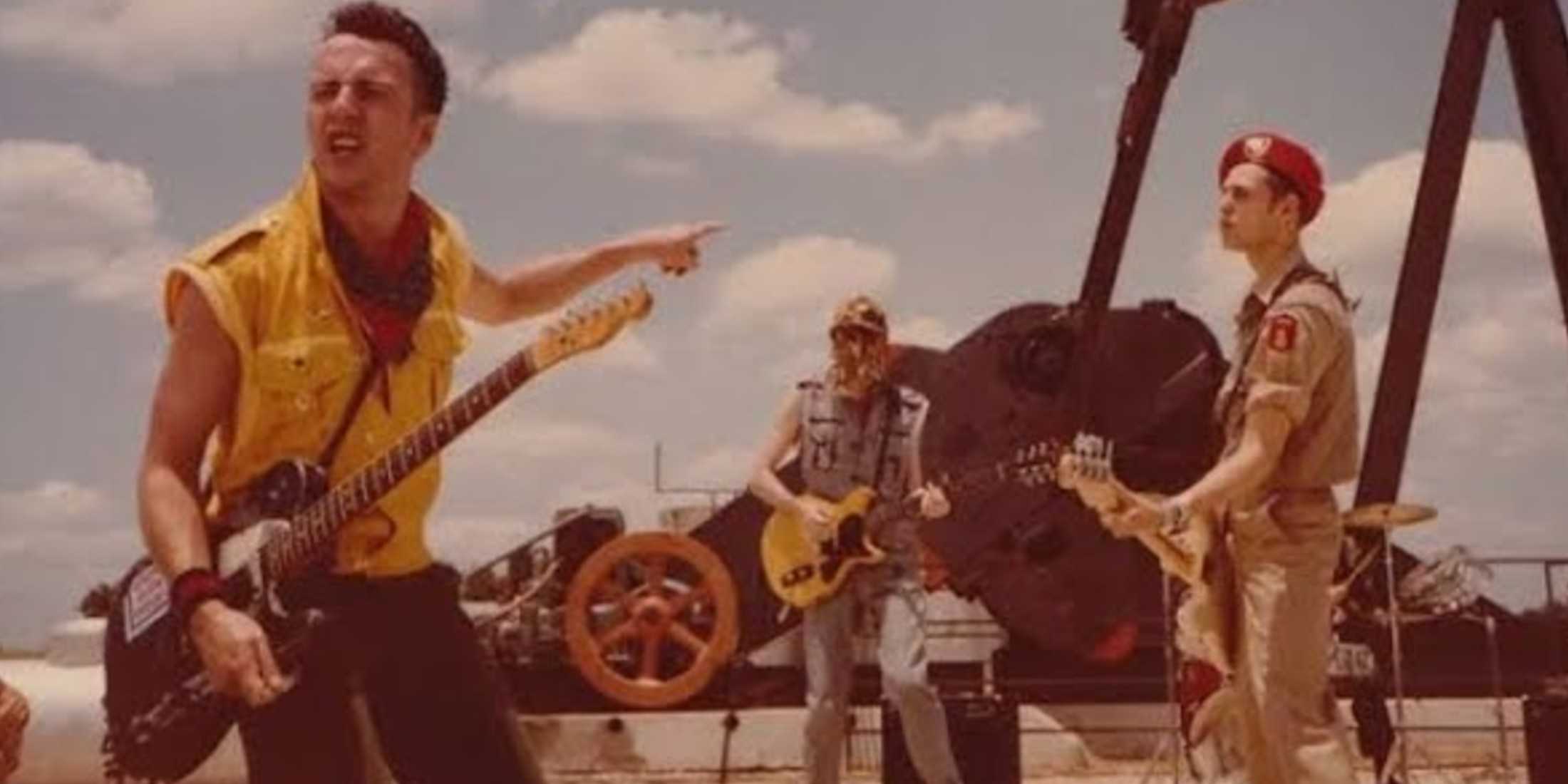 На заднем плане - голубое небо с облаками. За группой находится механический механизм. Слева, один участник носит черные брюки, желтую куртку и держит гитару. В центре, один участник - джинсы и джинсовую куртку, играет на гитаре. Участник справа - брюки, рубашка на пуговицах, шляпу и держит бас-гитару. Его тело смещено вправо.