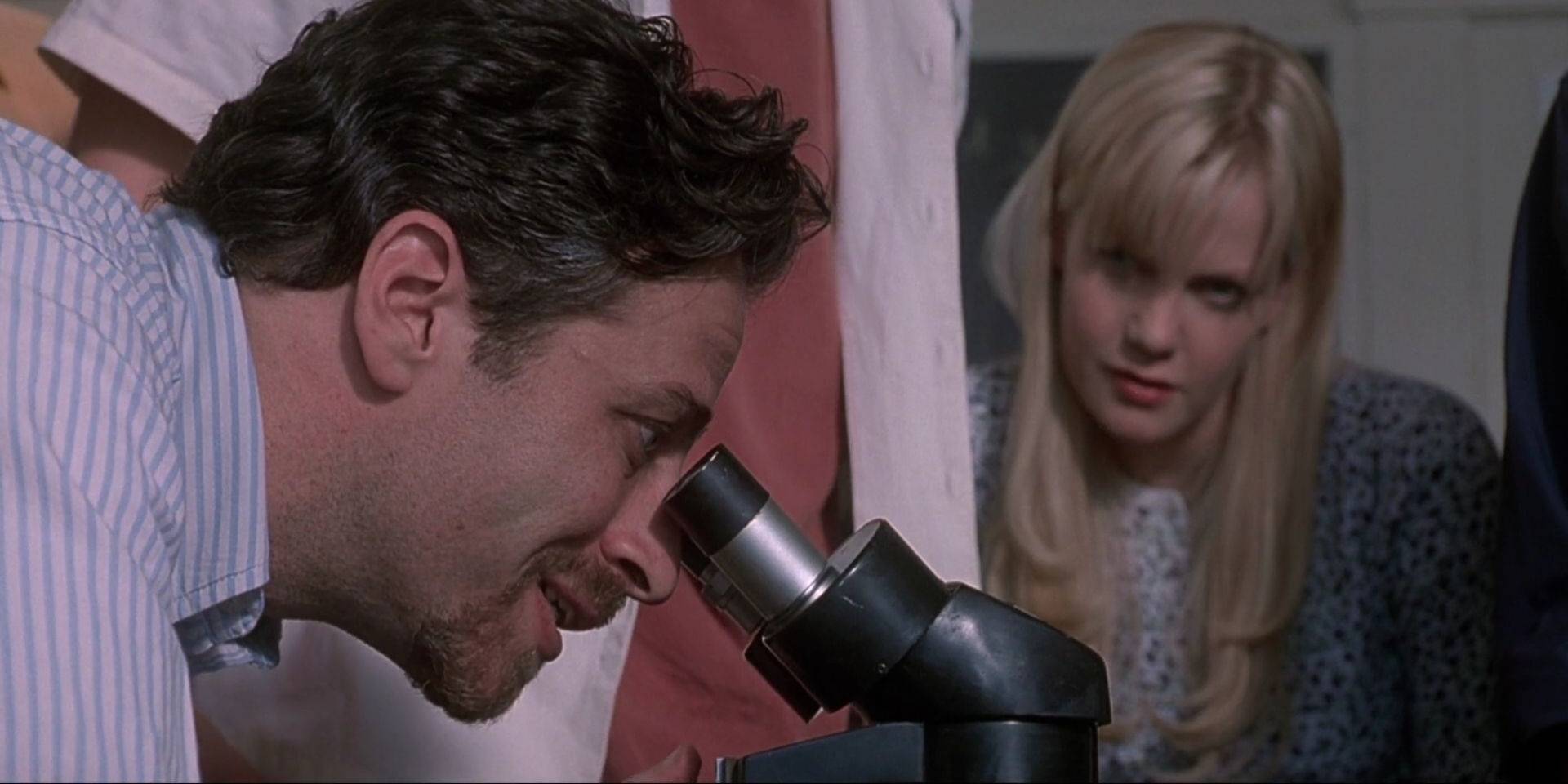 Джон Стюарт, в роли Фулронга, рассматривает под микроскопом, в то время как Мэрибет, сыгранная Лаурой Харрис, наблюдает за ним в фильме "The Faculty"